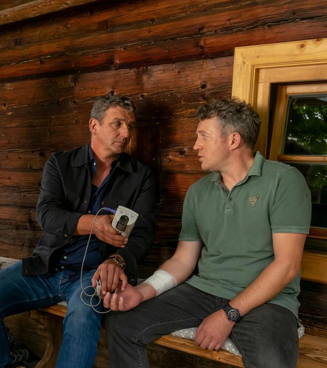 "Der Bergdoktor: Hochspannung": Vor seinem Haus sitzt Franz Herford (Josef Heynert) neben Dr. Martin Gruber (Hans Sigl), der ihm den Puls misst. Am Arm trägt Franz ein Verband. Neben ihnen steht Martha Herford (Anne Weinknecht) und blickt ihren Mann ängstlich an.