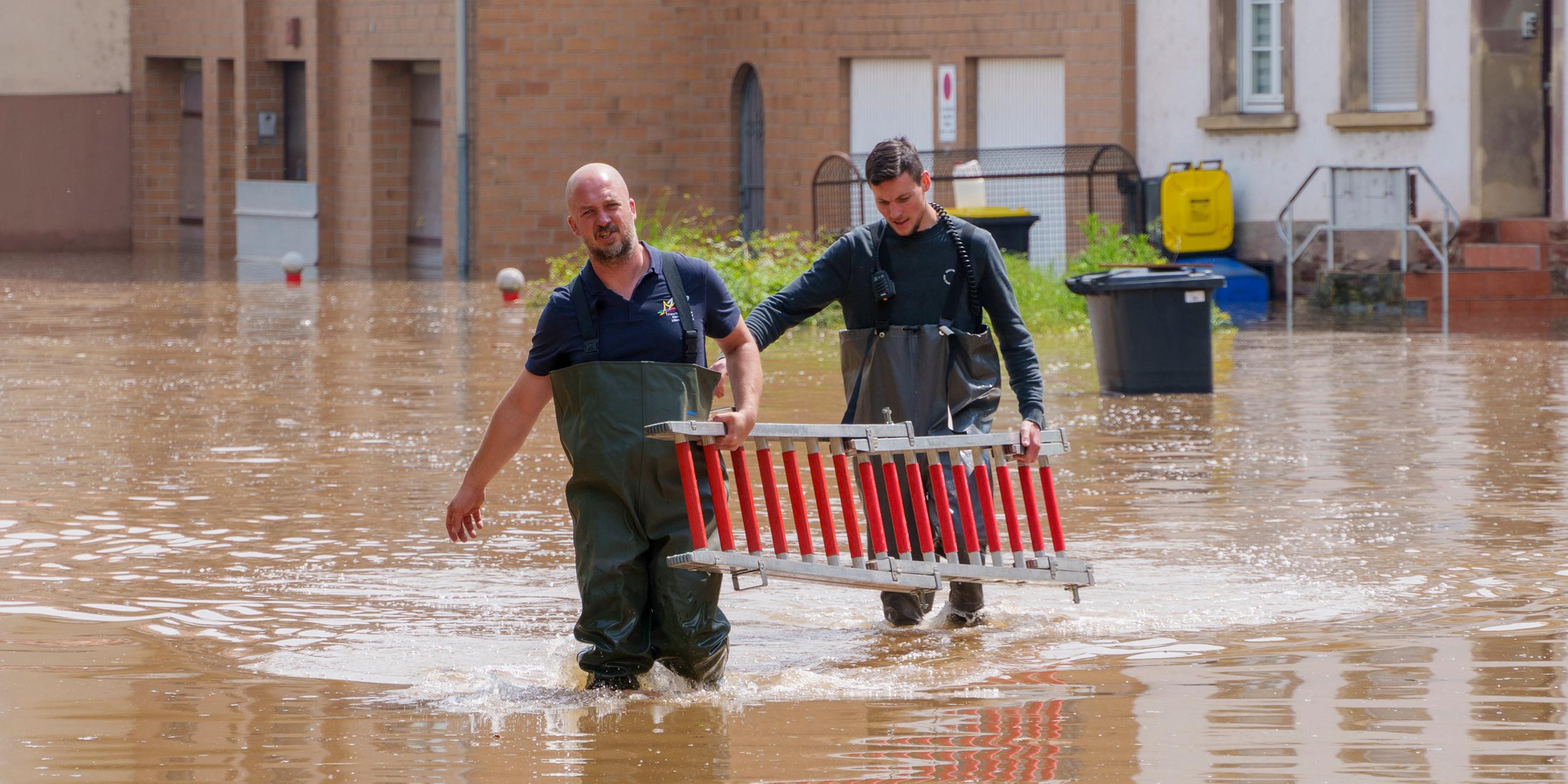 Einsatzkräfte der Freiwilligen Feuerwehr gehen mit einer Leiter durch eine überschwemmte Straße in Kleinblittersdorf im Saarland.