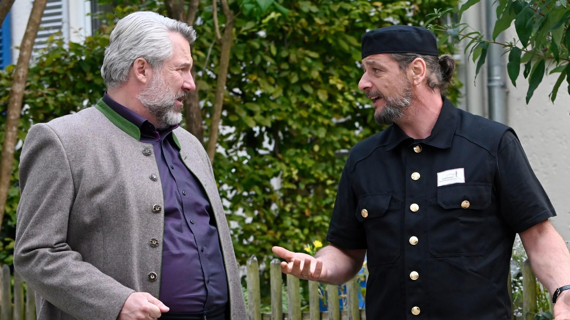 Kommissar Stadler (Dieter Fischer) und Quirin Lachner (Gerhard Greiner) stehen an einem Holzzaun und unterhalten sich angeregt. Lachner trägt schwarze kurzärmlige Arbeitskleidung und eine schwarze Kappe.