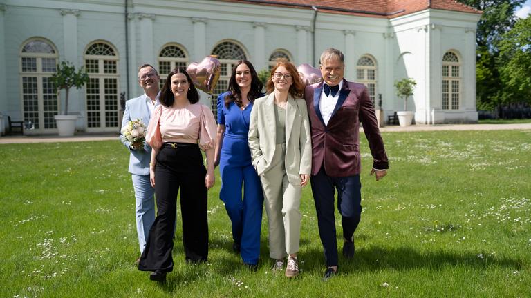 "Duell der Hochzeitsprofis": Marco Fuß, Amelie Borges, Madeleine Bolz, Marie Mika und Froonck laufen lächelnd über eine Wiese auf die Kamera zu. Im Hintergrund liegt die Orangerie in Potsdam.