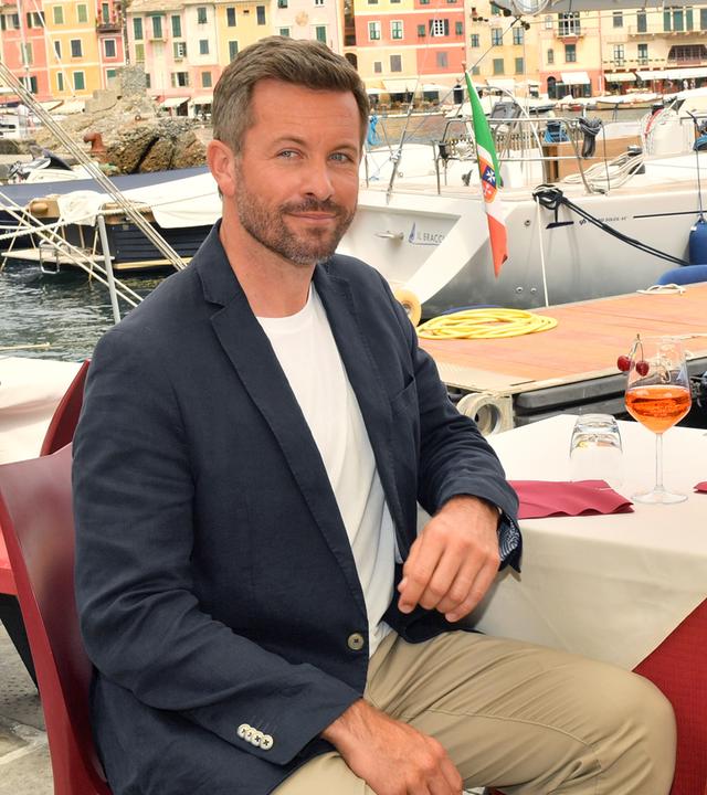 Sofie (Rebecca Dyson Smith) und Tom (Jan Hartmann) sitzen an einem Restauranttisch am Hafen und unterhalten sich. Im Hintergrund liegen Segelboote.