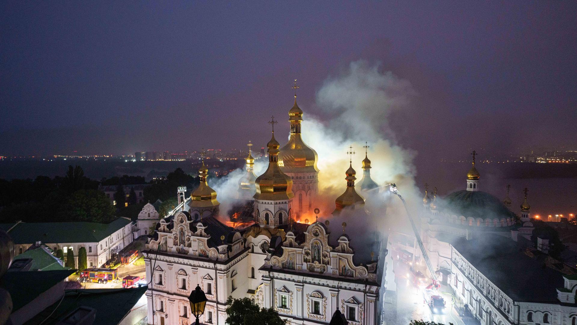 Eine große alte Klosteranlage bestehend aus einem weißen Gebäudekomplex mit goldenen Türmen wird von Feuerschein im Inneren erleuchtet und ist in den Nebel von Löschwasser gehüllt. Rechts und links vom Kloster in den Straßen sind ganz klein Feuerwehrfahrzeuge und Männer mit einer hohen Leiter und Löschschläuchen zu sehen.