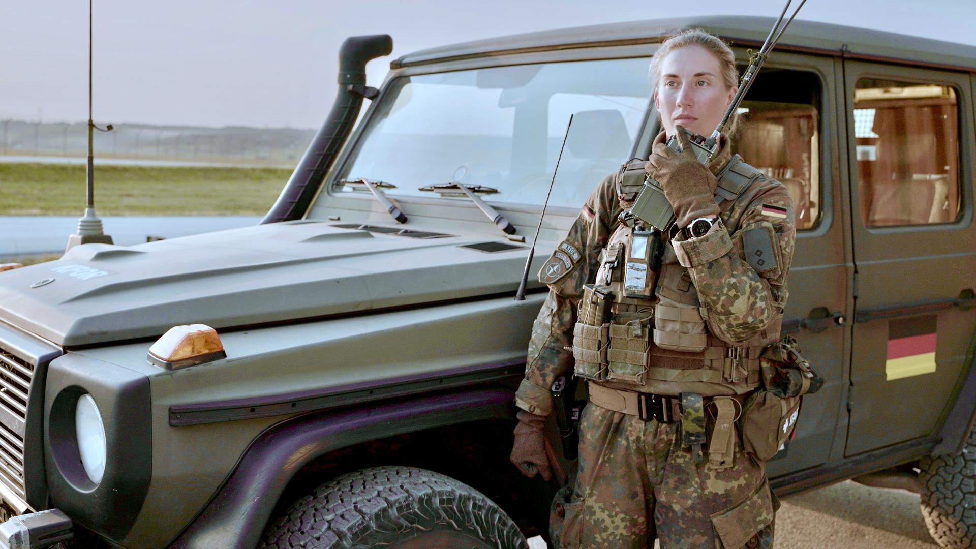 Alina steht in Flecktarn-Uniform und Schutzweste vor einem Bundeswehrauto. Sie hält das Handmikrofon eines Funkgeräts dicht an den Mund und spricht hinein.
