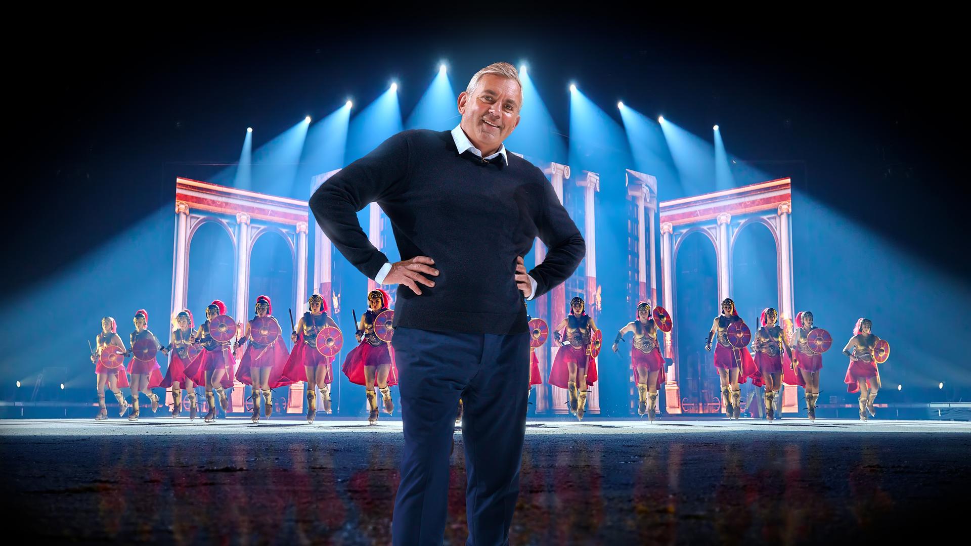 Das Bild zeigt eine Szene, die wie eine Bühnenperformance aussieht. Im Vordergrund steht ein Mann in einem dunkelblauen Pullover und einer blauen Hose. Er hat graue Haare und lächelt in die Kamera, wobei er seine Hände in die Hüften gestemmt hat. Hinter ihm sind eine Gruppe von Einkunstläufer:innen in Kostümen zu sehen, die wie römische Soldaten gekleidet sind. Sie tragen rote Umhänge, goldene Brustpanzer und Helme sowie rote Schilde, dazu Schlittschuhe. Der Hintergrund der Bühne hat eine Kulisse, die an römische Architektur erinnert, mit hohen Säulen und Bögen. Über ihnen sind blaue Lichter zu sehen, die auf die Bühne gerichtet sind und eine dramatische Beleuchtung erzeugen.