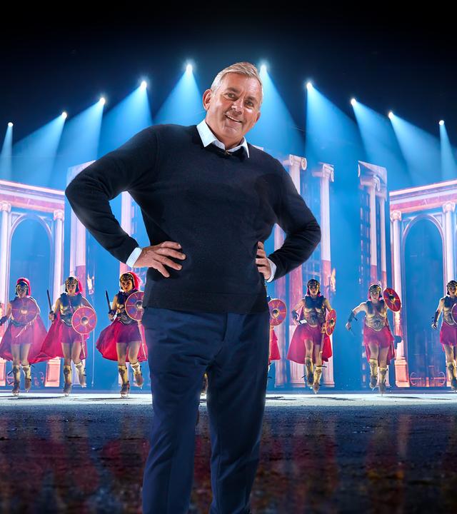 Das Bild zeigt eine Szene, die wie eine Bühnenperformance aussieht. Im Vordergrund steht ein Mann in einem dunkelblauen Pullover und einer blauen Hose. Er hat graue Haare und lächelt in die Kamera, wobei er seine Hände in die Hüften gestemmt hat. Hinter ihm sind eine Gruppe von Einkunstläufer:innen in Kostümen zu sehen, die wie römische Soldaten gekleidet sind. Sie tragen rote Umhänge, goldene Brustpanzer und Helme sowie rote Schilde, dazu Schlittschuhe. Der Hintergrund der Bühne hat eine Kulisse, die an römische Architektur erinnert, mit hohen Säulen und Bögen. Über ihnen sind blaue Lichter zu sehen, die auf die Bühne gerichtet sind und eine dramatische Beleuchtung erzeugen.