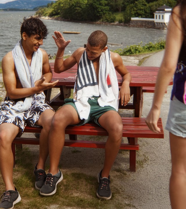 "After Benjamin: Holmlia For Life": Benjamin (Emil Sesay Stenseth) und Elias (Sam Ashraf) sitzen in Badekleidung am Osloer Meeresufer vor einem roten Holztisch auf einer Bank und klatschen sich ab. Sie wirken entspannt und gut gelaunt.