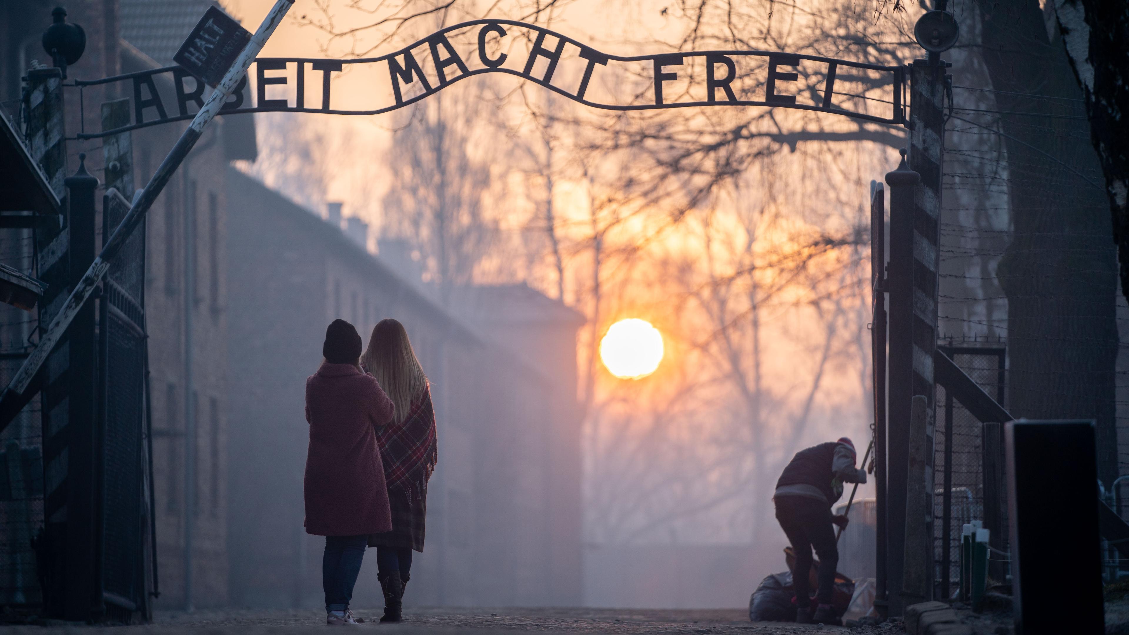Zwei Personen stehen vor dem Schriftzug "Arbeit macht frei" im KZ Auschwitz