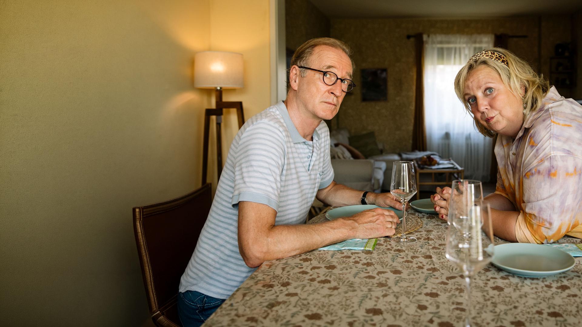 "I don't work here - Homeoffice": Heidi (Gabriela Maria Schmeide) und Walter (Peter Lohmeyer) sitzen sich am gedeckten Tisch gegenüber. Heidi hat die Unterarme auf dem Tisch abgestützt, lehnt sich nach vorne und schaut zu ihrer linken Seite. Auch Walter blickt durch seine runde Brille etwas ängstlich zur Seite und ist dabei leicht nach vorne geneigt. Seinen hellblauen Teller hält er an den Seiten fest.
