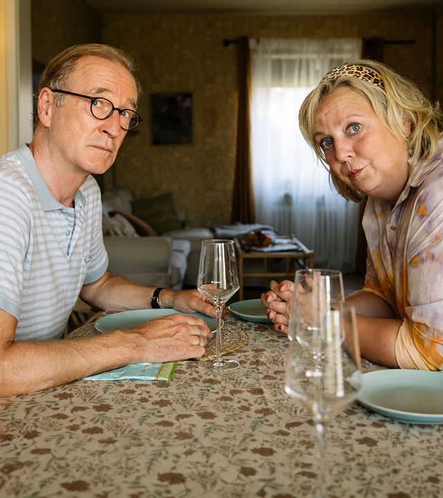 "I don't work here - Homeoffice": Heidi (Gabriela Maria Schmeide) und Walter (Peter Lohmeyer) sitzen sich am gedeckten Tisch gegenüber. Heidi hat die Unterarme auf dem Tisch abgestützt, lehnt sich nach vorne und schaut zu ihrer linken Seite. Auch Walter blickt durch seine runde Brille etwas ängstlich zur Seite und ist dabei leicht nach vorne geneigt. Seinen hellblauen Teller hält er an den Seiten fest.