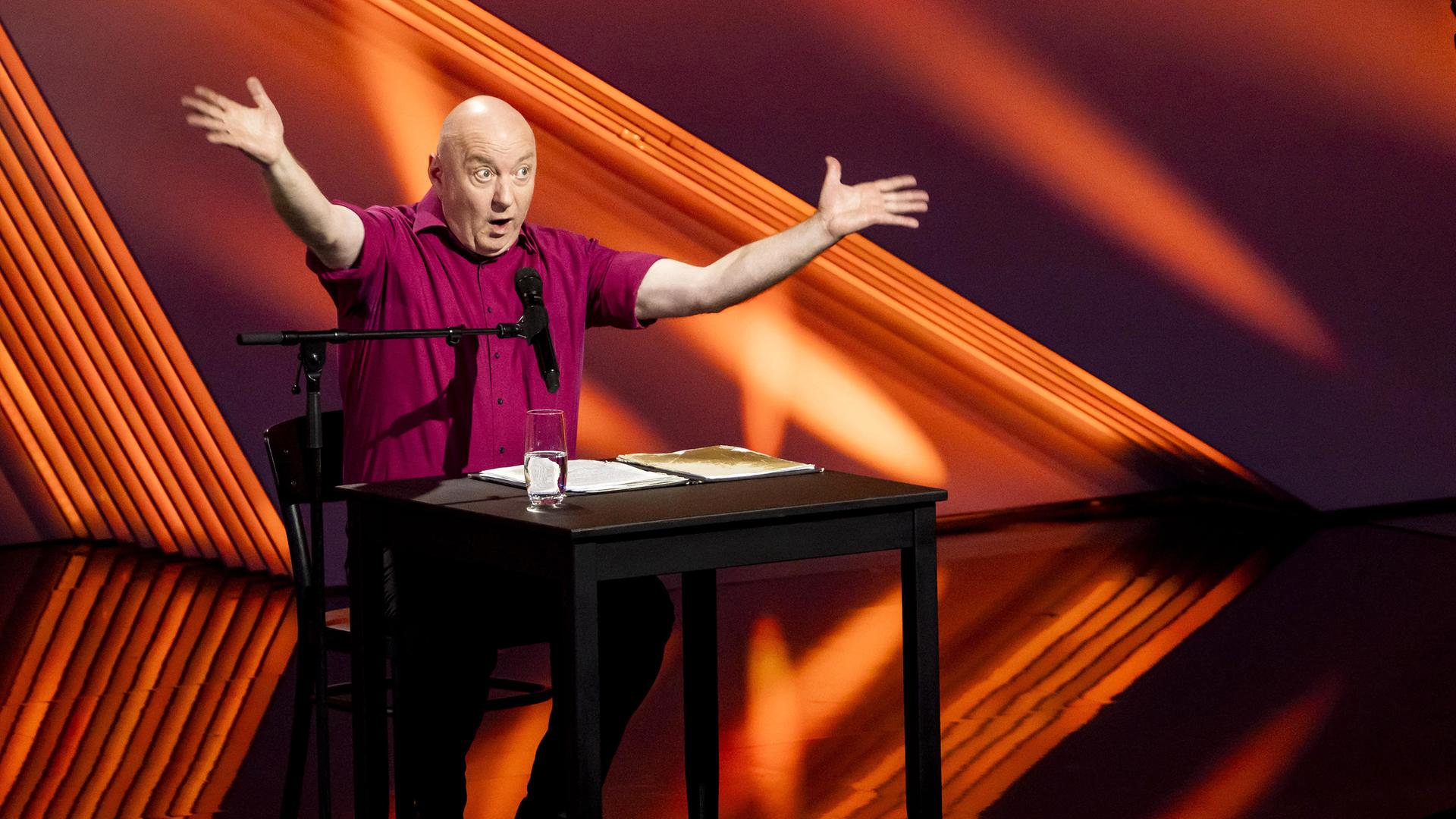 Horst Evers sitzt auf der linken Bildseite an einem Tisch. Er hebt beide Arme an der Seite. Der Hintergrund ist rot beleuchtet.  