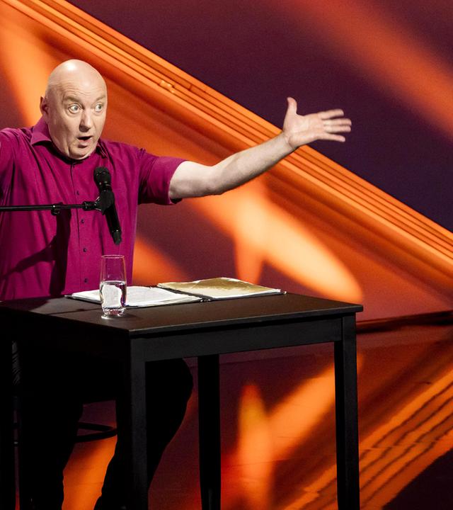 Horst Evers sitzt auf der linken Bildseite an einem Tisch. Er hebt beide Arme an der Seite. Der Hintergrund ist rot beleuchtet.  