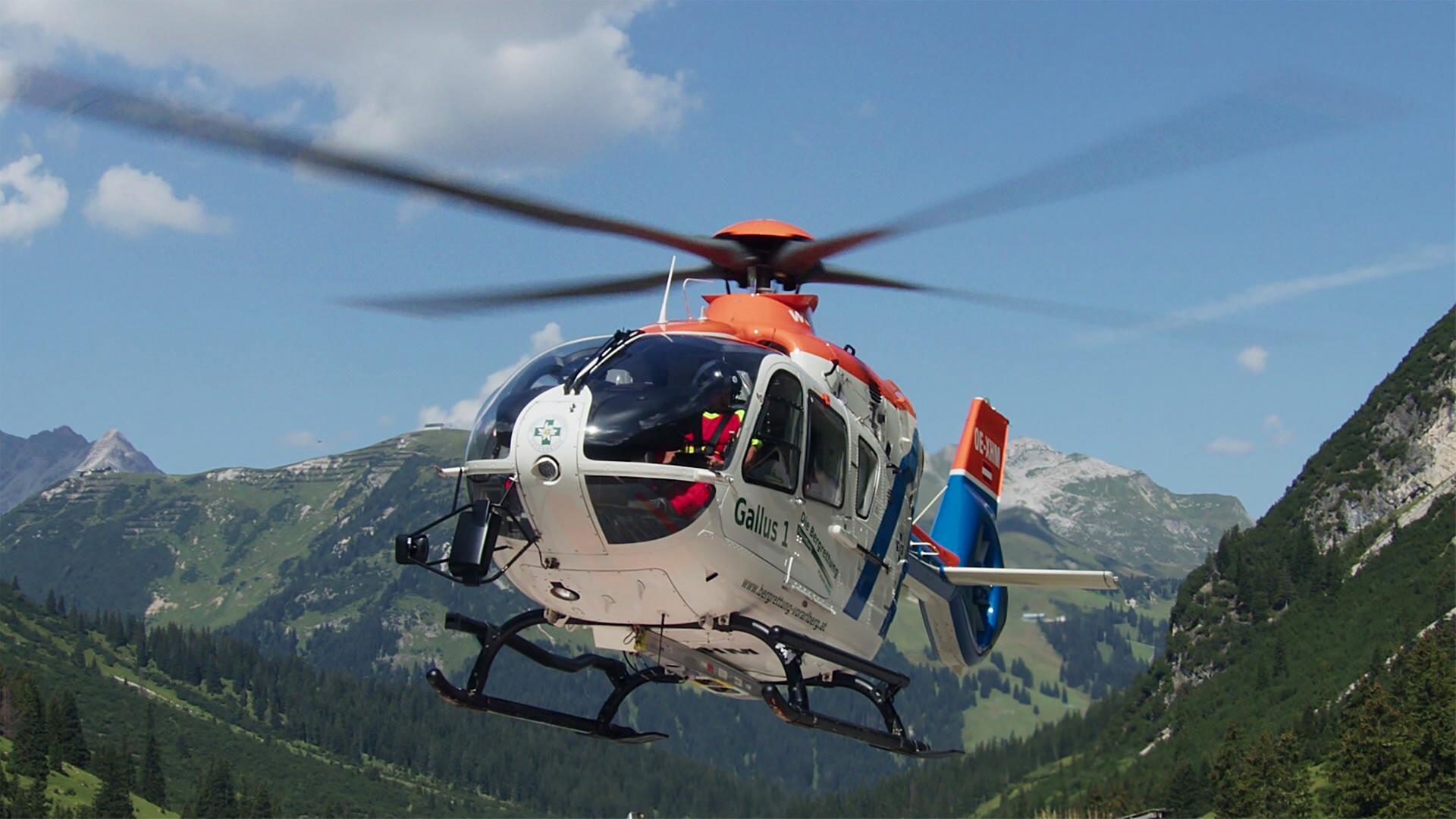 Das Bild zeigt einen Hubschrauber in der Luft, umgeben von einem malerischen Bergpanorama. Der Hubschrauber hat eine weiße Hauptkarosserie mit orangefarbenen und blauen Akzenten. Die Rotorblätter sind in Bewegung und erzeugen das typische Geräusch des Fliegens. Im Hintergrund sind grüne Berge und bewaldete Flächen zu sehen, während der Himmel strahlend blau ist mit einigen kleinen, weißen Wolken. Die Szene vermittelt ein Gefühl von Weite und Freiheit, das mit der Faszination für Hubschrauber verbunden ist.