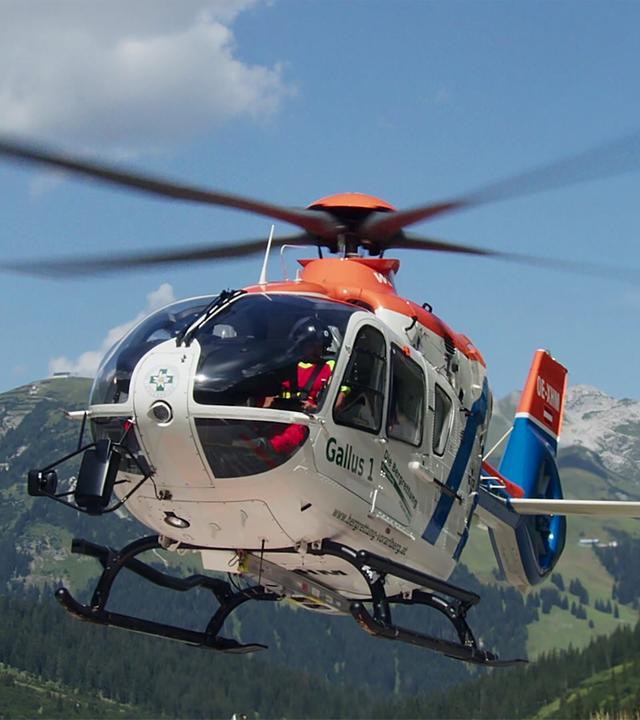 Das Bild zeigt einen Hubschrauber in der Luft, umgeben von einem malerischen Bergpanorama. Der Hubschrauber hat eine weiße Hauptkarosserie mit orangefarbenen und blauen Akzenten. Die Rotorblätter sind in Bewegung und erzeugen das typische Geräusch des Fliegens. Im Hintergrund sind grüne Berge und bewaldete Flächen zu sehen, während der Himmel strahlend blau ist mit einigen kleinen, weißen Wolken. Die Szene vermittelt ein Gefühl von Weite und Freiheit, das mit der Faszination für Hubschrauber verbunden ist.