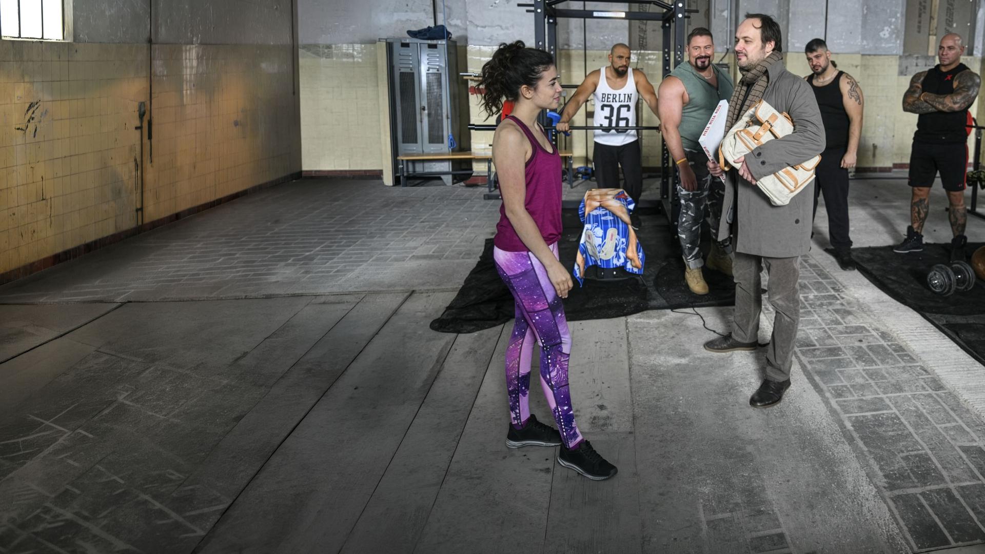 "Hüftkreisen mit Nancy": Nancy (Helen Woigk) und Max (Felix Knopp) stehen sich vor den Geräten des Fitnessstudios gegenüber und werden dabei von den anderen Mitgliedern beobachtet.