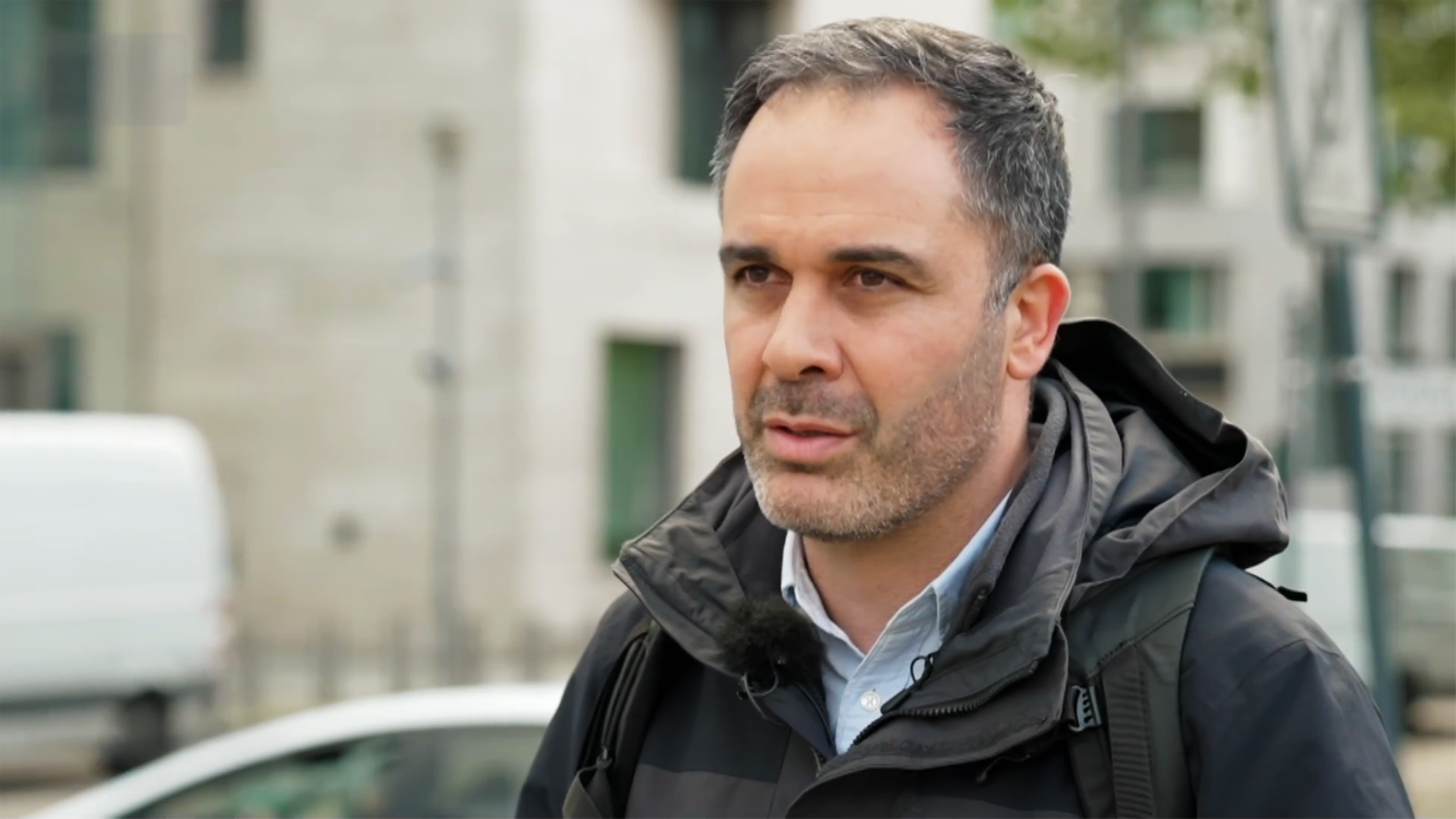 Hüseyin Doğru, ein Mann mittleren Alters mit Dreitagebart und kurzen, leicht angegrauten Haaren trägt eine dunkle Jacke über einem Hemd und steht irgendwo in Berlin auf der Straße.