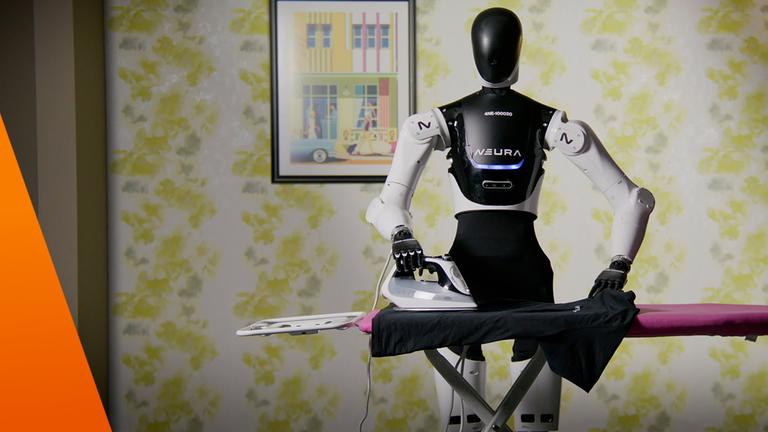 Ein schwarz-weißer humanoider Roboter steht vor einem Bügelbrett mit pinkfarbenem Bezug. Eine Hand liegt auf dem Griff eines Bügeleisens, die andere auf einem dunklen Kleidungsstück, das über das Brett ausgebreitet ist. Auf der Brust des Roboters steht „NEURA“. Im Hintergrund sind eine geblümte Tapete, ein gerahmtes Bild und eine Stehlampe zu sehen.
