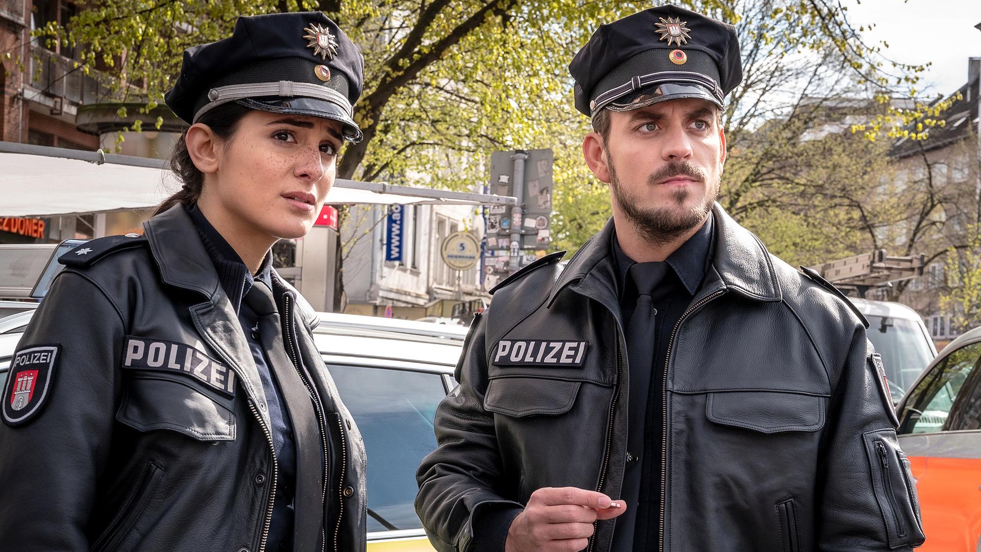  Die Polizistin Pinar Aslan (Aybi Era) und ihr Kollege Kris Freiberg (Marc Barthel) stehen nebeneinander und blicken in die gleiche Richtung. Hinter ihnen stehen ein Streifen- und ein Notarztwagen.