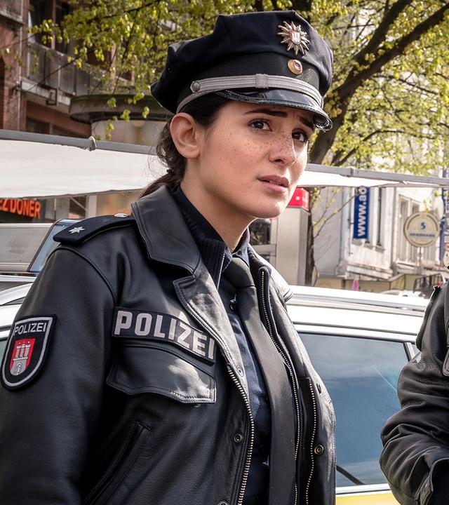  Die Polizistin Pinar Aslan (Aybi Era) und ihr Kollege Kris Freiberg (Marc Barthel) stehen nebeneinander und blicken in die gleiche Richtung. Hinter ihnen stehen ein Streifen- und ein Notarztwagen.