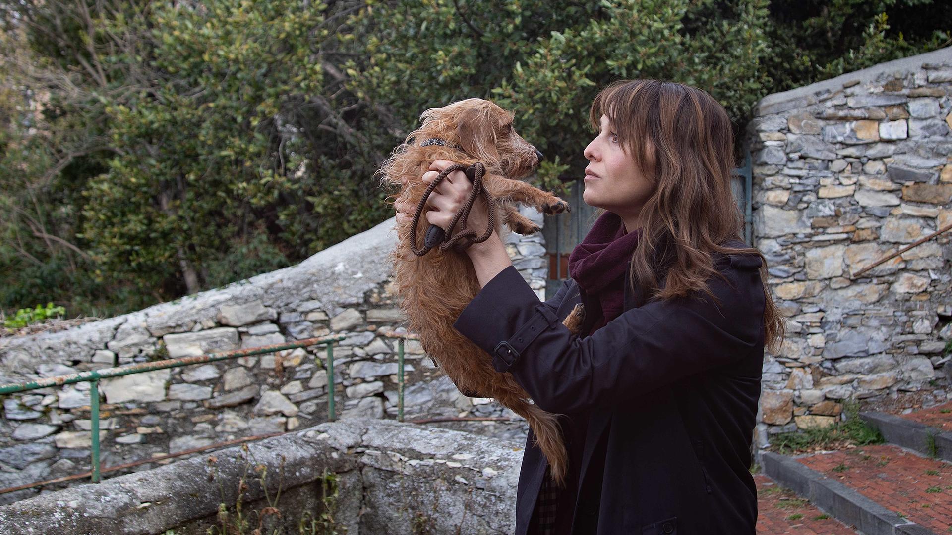 Petra (Paola Cortellesi) steht draußen auf einer großen Steintreppe, hält mit beiden Händen einen kleinen Hund hoch und schaut ihn liebevoll an.