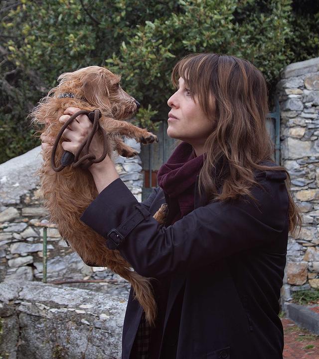 Petra (Paola Cortellesi) steht draußen auf einer großen Steintreppe, hält mit beiden Händen einen kleinen Hund hoch und schaut ihn liebevoll an.
