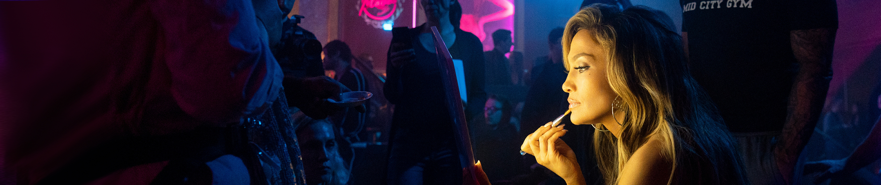 Backstage im Stripclub sitzt Ramona (Jennifer Lopez) an einem Tisch und schminkt sich für ihren Auftritt. Im restlichen dunkleren Raum sieht man andere Frauen und ein großer Mann steht direkt hinter Ramona.