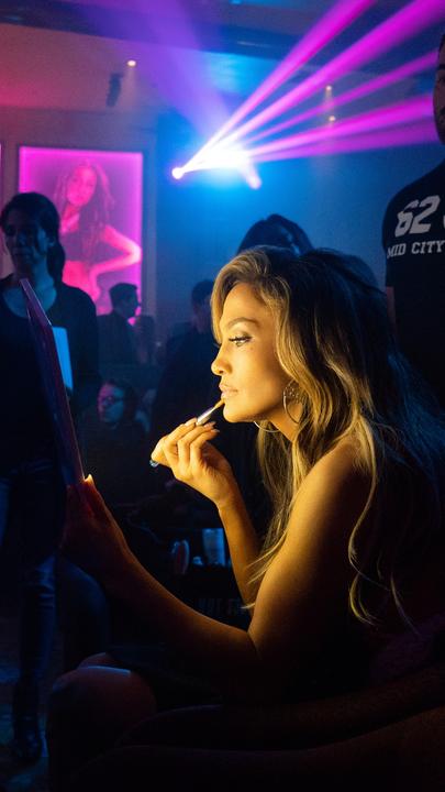 Backstage im Stripclub sitzt Ramona (Jennifer Lopez) an einem Tisch und schminkt sich für ihren Auftritt. Im restlichen dunkleren Raum sieht man andere Frauen und ein großer Mann steht direkt hinter Ramona.