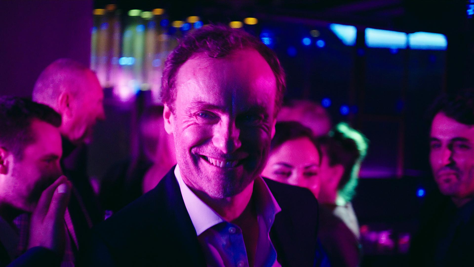 "I AM THE GREATEST - Agent of Change": Karsten Drechsler (Mark Waschke) steht in einer Bar und lächelt gewinnend.