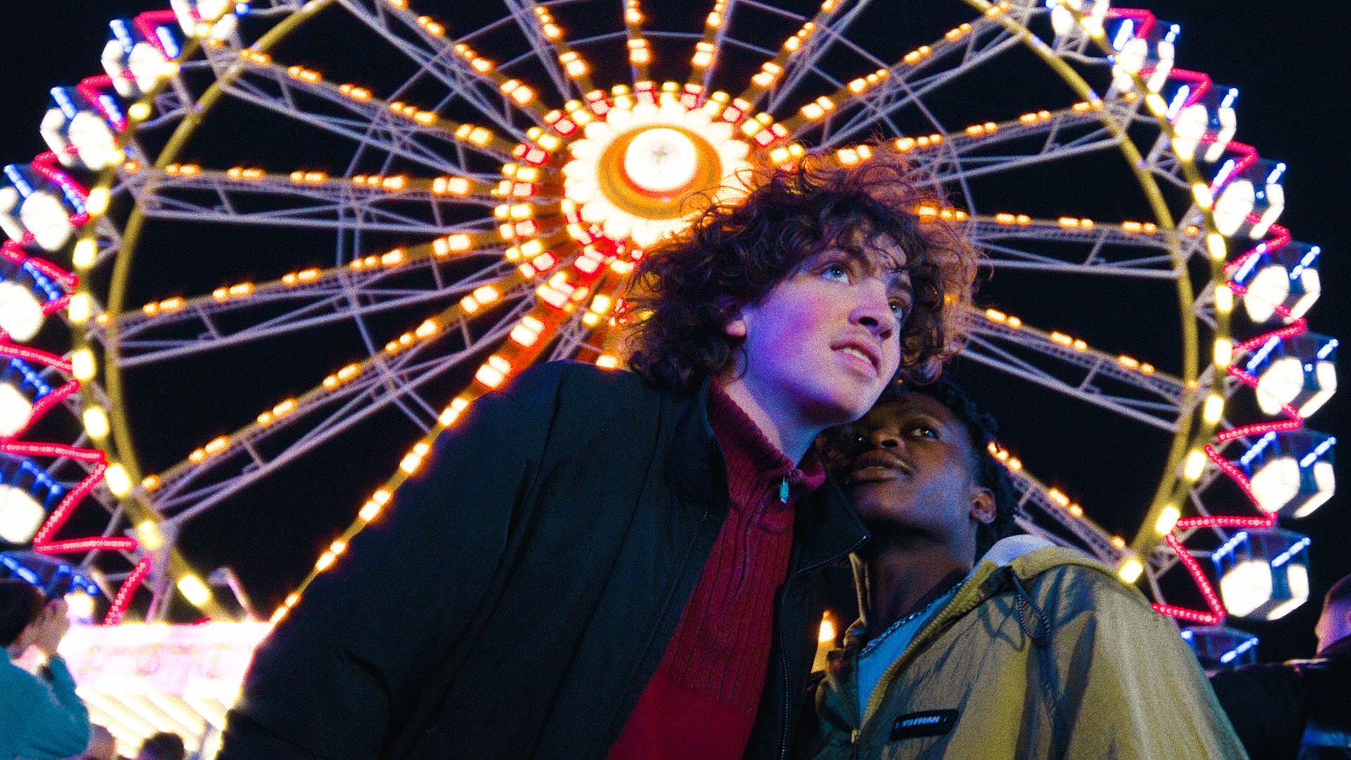 "I AM THE GREATEST - I am the Greatest": Lenny (Carlo Schmitt) und Mo (Thapelo Mashiane) verbringen zusammen ein Date auf dem Jahrmarkt. Im Hintergrund sieht man ein leuchtendes Riesenrad.
