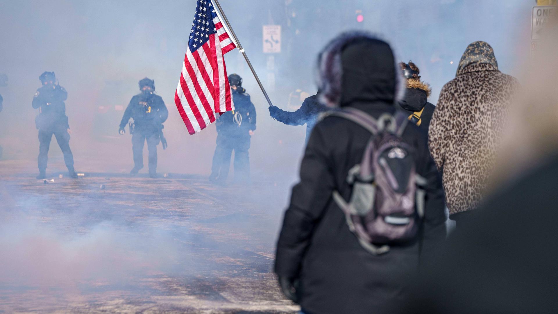 Mit dem Rücken zum Betrachter stehen demonstrierende Menschen mit einer US-Flagge bewaffneten Mitgliedern der ICE-Beamten gegenüber.