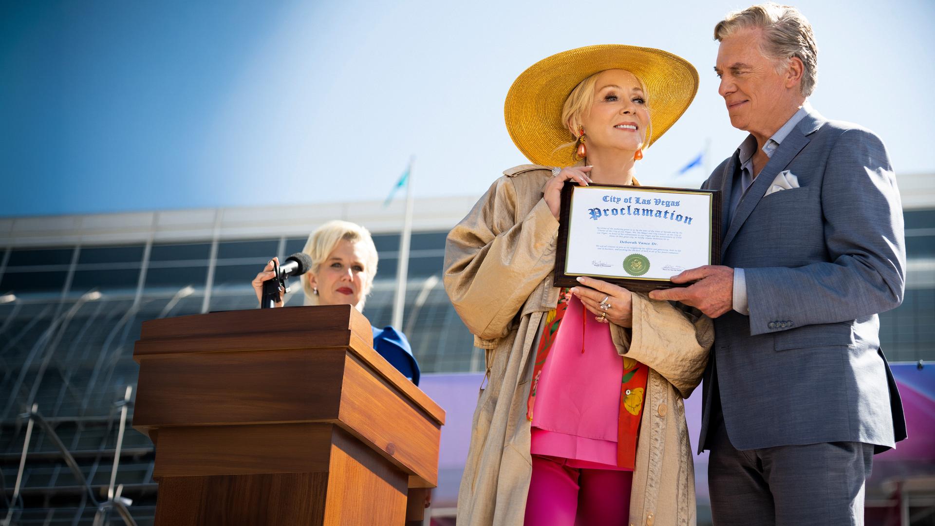 "Hacks - Ich denke, sie wird": Deborah (Jean Smart) hält lächelnd eine gerahmte Urkunde, während Marty (Christopher McDonald) neben ihr steht, das Dokument ebenfalls hält und sie dabei ansieht. Im Hintergrund steht Bürgermeisterin Jo (Lauren Weedman) an einem Rednerpult und beobachtet das Geschehen.