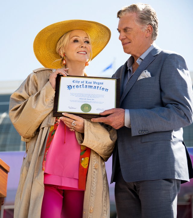 "Hacks - Ich denke, sie wird": Deborah (Jean Smart) hält lächelnd eine gerahmte Urkunde, während Marty (Christopher McDonald) neben ihr steht, das Dokument ebenfalls hält und sie dabei ansieht. Im Hintergrund steht Bürgermeisterin Jo (Lauren Weedman) an einem Rednerpult und beobachtet das Geschehen.