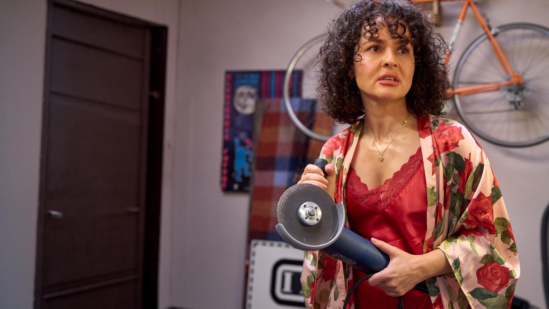 "Ich dich auch! - Der Stichtag": Jackie (Nagmeh Alaei) steht im Spitzennachthemd völlig aufgelöst im Raum. Sie hält eine Schneidemaschine in den Händen.