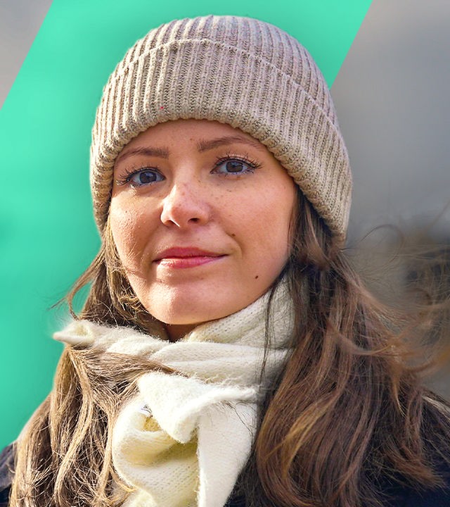 Eine junge Frau schaut fröhlich nach vorne. Sie trägt eine Mütze und einen weißen Schal.