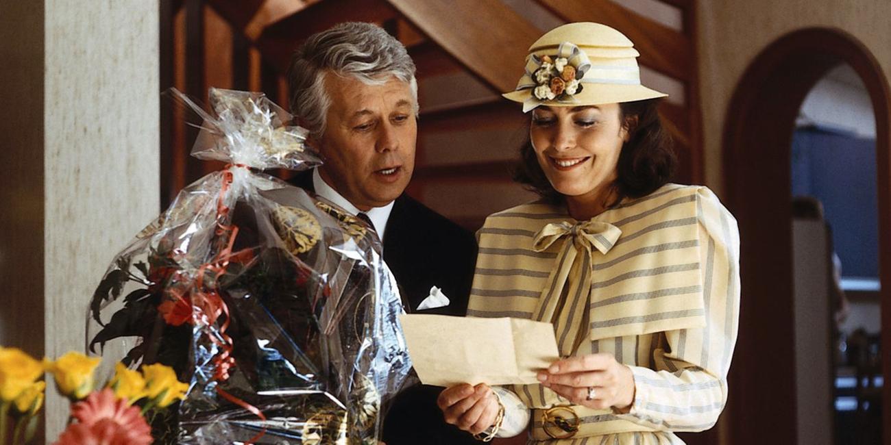 Werner Schumann und Angelika Graf stehen in festlicher Kleidung vor lauter Geschenkkörben mit Blumen darin. Angelika blickt lächelnd auf einen Brief, den sie in beiden Händen hält. Werner neigt sich zu ihr herüber und blickt ebenfalls interessiert hinunter zu dem Brief in Angelikas Händen.