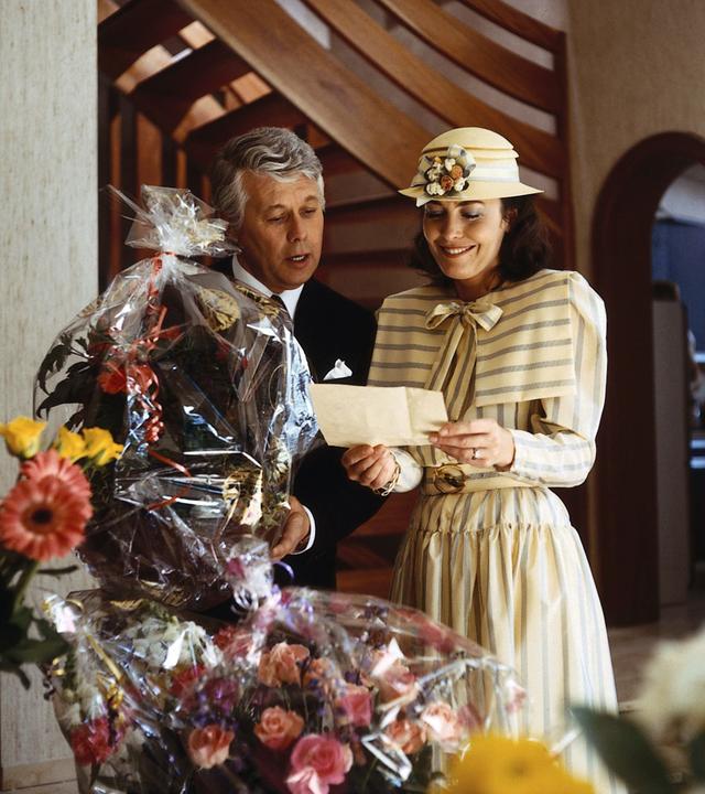 Werner Schumann und Angelika Graf stehen in festlicher Kleidung vor lauter Geschenkkörben mit Blumen darin. Angelika blickt lächelnd auf einen Brief, den sie in beiden Händen hält. Werner neigt sich zu ihr herüber und blickt ebenfalls interessiert hinunter zu dem Brief in Angelikas Händen.