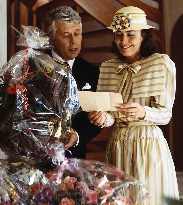 Werner Schumann und Angelika Graf stehen in festlicher Kleidung vor lauter Geschenkkörben mit Blumen darin. Angelika blickt lächelnd auf einen Brief, den sie in beiden Händen hält. Werner neigt sich zu ihr herüber und blickt ebenfalls interessiert hinunter zu dem Brief in Angelikas Händen.