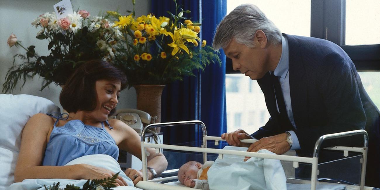 Angi liegt in einem Krankenhausbett und blickt lächelnd auf ihr Neugeborenes, das in einem Kinderbettchen liegt. Werner steht neben ihrem Bett und beugt sich fürsorglich über das Baby. Ringsherum stehen Blumen im Zimmer.