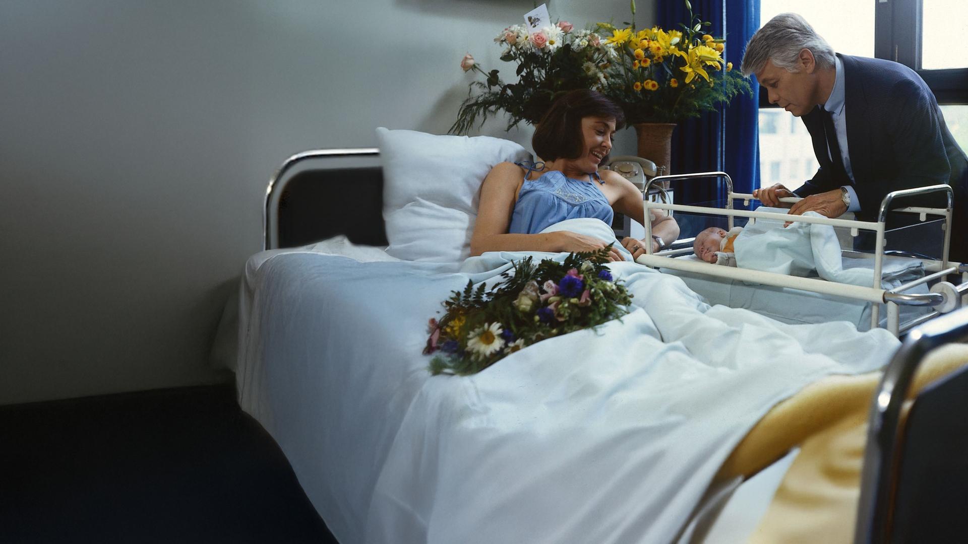 Angi liegt in einem Krankenhausbett und blickt lächelnd auf ihr Neugeborenes, das in einem Kinderbettchen liegt. Werner steht neben ihrem Bett und beugt sich fürsorglich über das Baby. Ringsherum stehen Blumen im Zimmer.