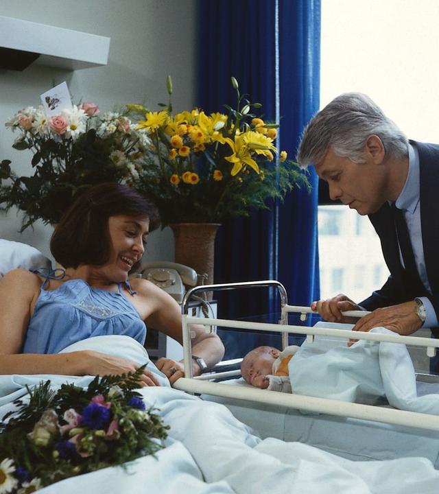 Angi liegt in einem Krankenhausbett und blickt lächelnd auf ihr Neugeborenes, das in einem Kinderbettchen liegt. Werner steht neben ihrem Bett und beugt sich fürsorglich über das Baby. Ringsherum stehen Blumen im Zimmer.