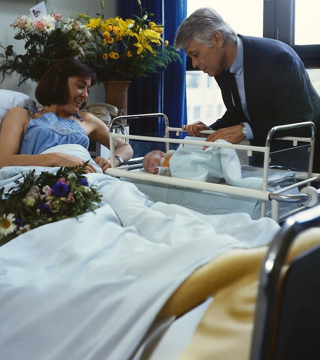 Angi liegt in einem Krankenhausbett und blickt lächelnd auf ihr Neugeborenes, das in einem Kinderbettchen liegt. Werner steht neben ihrem Bett und beugt sich fürsorglich über das Baby. Ringsherum stehen Blumen im Zimmer.