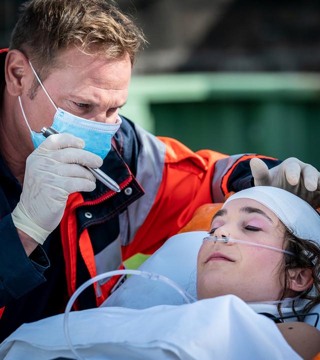 "Notruf Hafenkante - Ich liebe Dich": Großaufnahme von Dr. Haase (Fabian Harloff), der am Kopfende einer Patiententrage steht und mit der Pupillenleuchte in die Augen einer Patientin (Anni C. Salander) leuchtet.