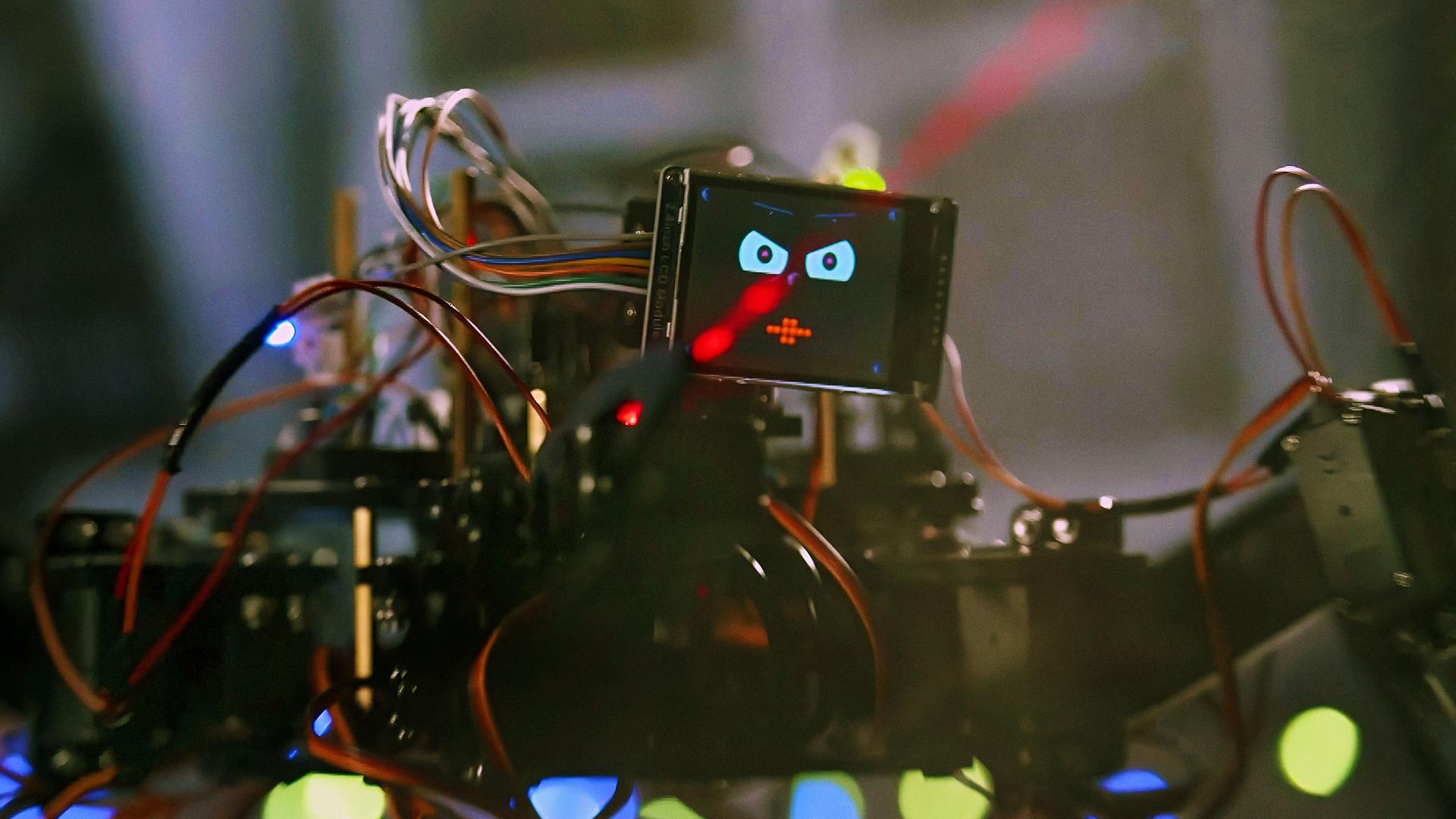 Ein spinnenartiger kleiner Roboter mit vielen Kabeln und Drähten sitzt vor einer Keyboard-Tastatur und drückt mit einem seiner Greifarme auf eine Taste. Als Kopf hat er einen kleinen Bildschirm, auf dem sein lachendes Gesicht zu sehen ist. Der Hintergrund ist unscharf, auf einem Bildschirm ist eine bunte Computeranimation zu erkennen.