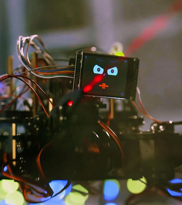 Ein spinnenartiger kleiner Roboter mit vielen Kabeln und Drähten sitzt vor einer Keyboard-Tastatur und drückt mit einem seiner Greifarme auf eine Taste. Als Kopf hat er einen kleinen Bildschirm, auf dem sein lachendes Gesicht zu sehen ist. Der Hintergrund ist unscharf, auf einem Bildschirm ist eine bunte Computeranimation zu erkennen.
