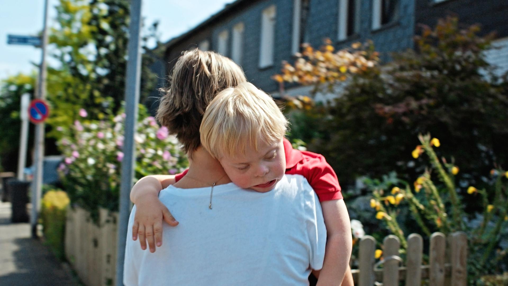 Rückansicht einer Frau in weißem T-Shirt, die einen kleinen schlafenden Jungen über die Schulter trägt. Im Hintergrund sieht man blühende Blumen in Vorgärten von Häusern.