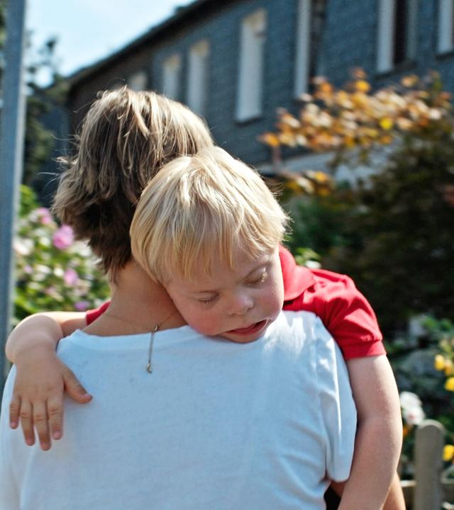 Rückansicht einer Frau in weißem T-Shirt, die einen kleinen schlafenden Jungen über die Schulter trägt. Im Hintergrund sieht man blühende Blumen in Vorgärten von Häusern.