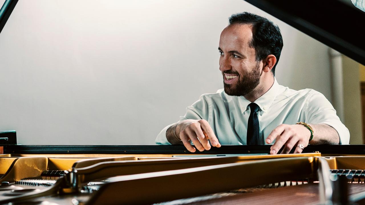 Blick durch den aufgeklappten Deckel des Flügels auf den am Instrument sitzenden Pianisten Igor Levit. Er blickt lächelnd zur Seite. Beide Arme liegen auf dem Instrument. Gekleidet ist er mit einem weißen Hemd und einer schwarzen Krawatte. Sein Gesicht spiegelt sich oben im Deckel des Flügels.