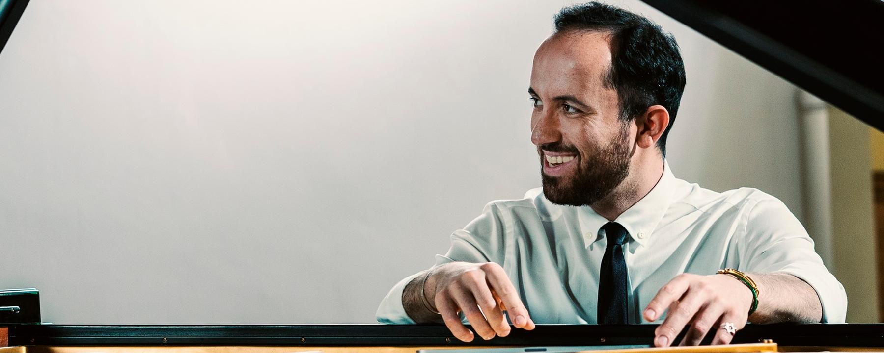 Blick durch den aufgeklappten Deckel des Flügels auf den am Instrument sitzenden Pianisten Igor Levit. Er blickt lächelnd zur Seite. Beide Arme liegen auf dem Instrument. Gekleidet ist er mit einem weißen Hemd und einer schwarzen Krawatte. Sein Gesicht spiegelt sich oben im Deckel des Flügels.