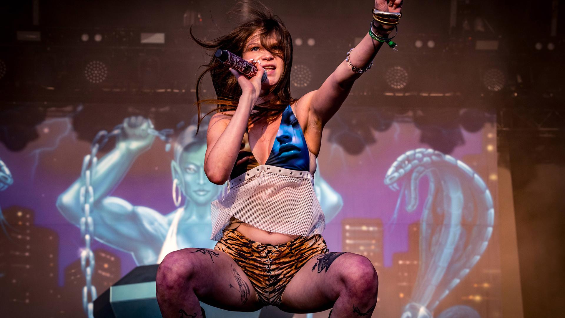 Eine leicht bekleidete Frau mit Tattoos auf den Beinen hockt auf einer Bühne uns singt in ein Mikro, während sie die linke Faust nach oben reckt.