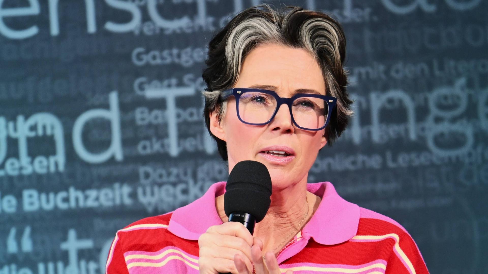 Eine Frau mittleren Alters mit kurzen dunklen Haaren, die vorne graue Strähnen haben, großer blauer Kunststoffbrille und pink gestreiftem Pullover, spricht in ein Mikrofon.