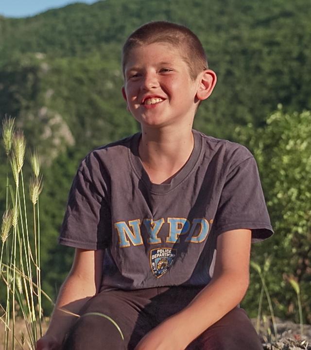 Ilija sitzt auf einem Felsen und lächelt fröhlich in die Kamera. Die Sonne scheint. Im Hintergrund kann man einen bewaldeten Hang erkennen. Alles wirkt sehr ruhig und friedlich.